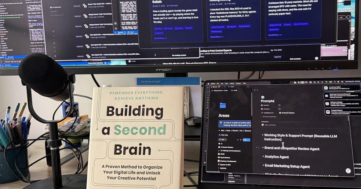 Tiago Forte's book Building a Second Brain propped on a working desk, beside a podcast mic, with a laptop showing the author's prompts workspace and a large monitor showing his blog in the background.